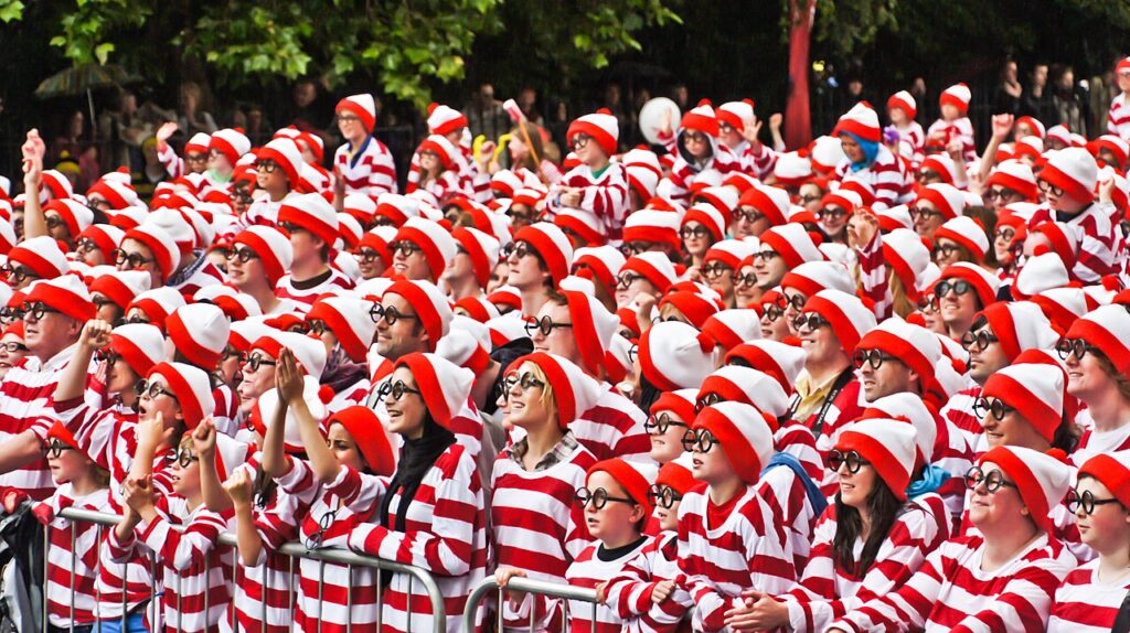 A large crowd of people dressed identically in red-and-white striped shirts, red-and-white bobble hats, and round black glasses, resembling the character "Wally" from the Where’s Wally? (known as Where’s Waldo? in the U.S.) book series. The participants are gathered behind a metal barrier in an outdoor setting under green trees, many smiling and gesturing enthusiastically. This event was part of an attempt to set a world record for the largest gathering of people dressed as Wally. Photographer: William Murphy License: Creative Commons Attribution-Share Alike 2.0 Generic Source URL: https://commons.wikimedia.org/wiki/File:Where%E2%80%99s_Wally_World_Record_(5846729480).jpg