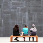 Three visitors to an art exhibition gallery or museum sitting on a bench with their backs to us, facing a wall-sized painting or mural. The artwork is a dark slate surface brushed with distressed strokes in irregularly rectangular patches all across its surface